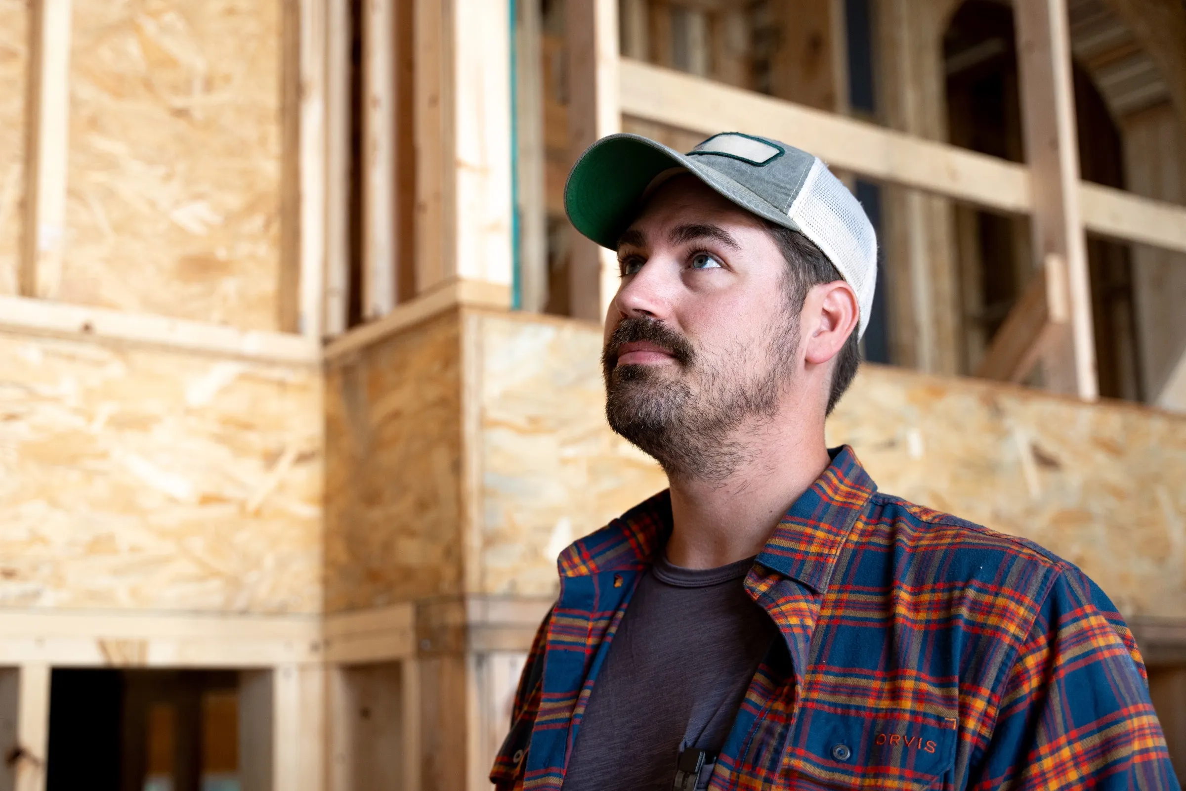 A builder in a cap and plaid shirt looks up inside a timber-framed house