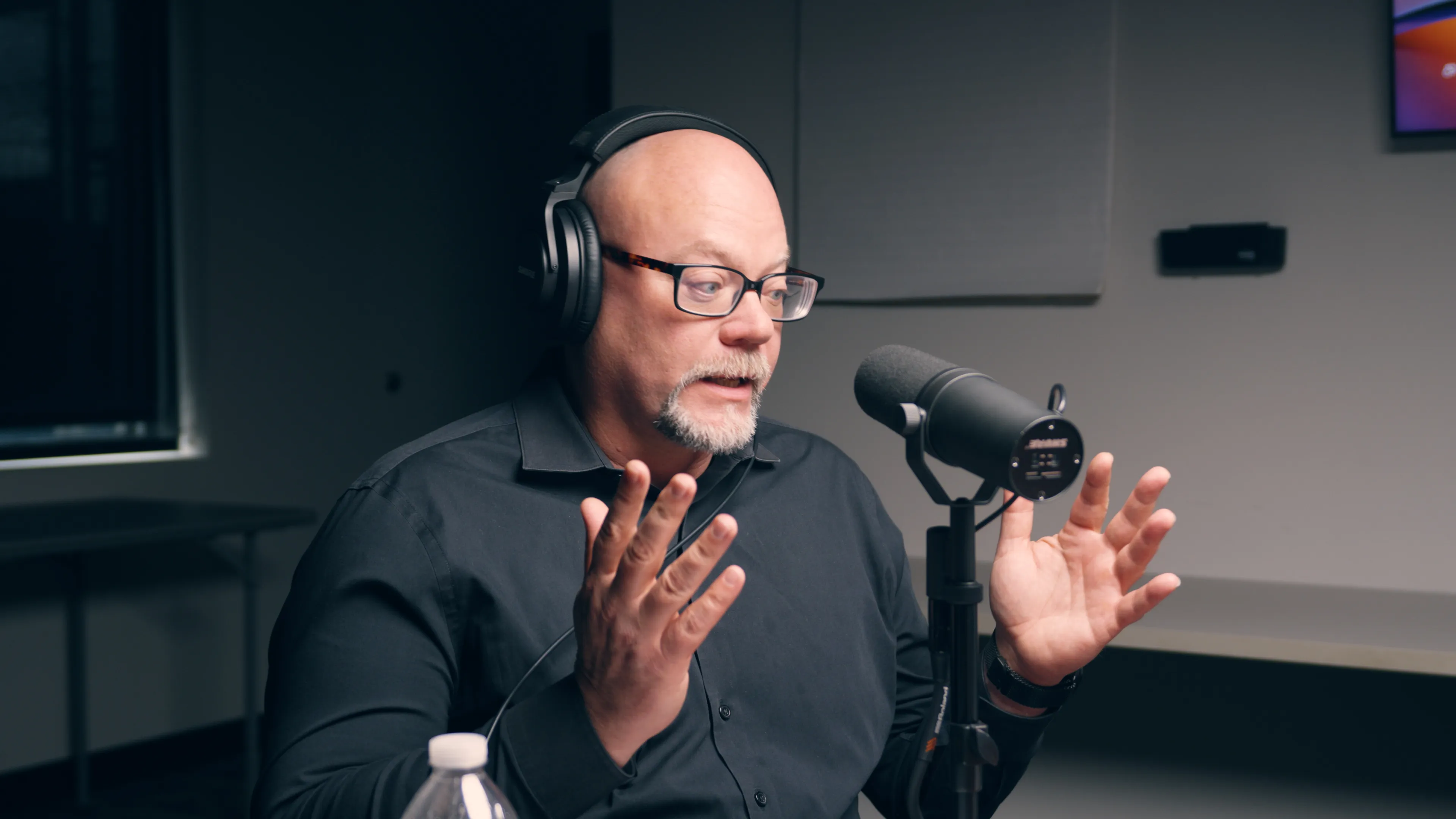 A man in headphones talks with both hands raised at a podcast microphone
