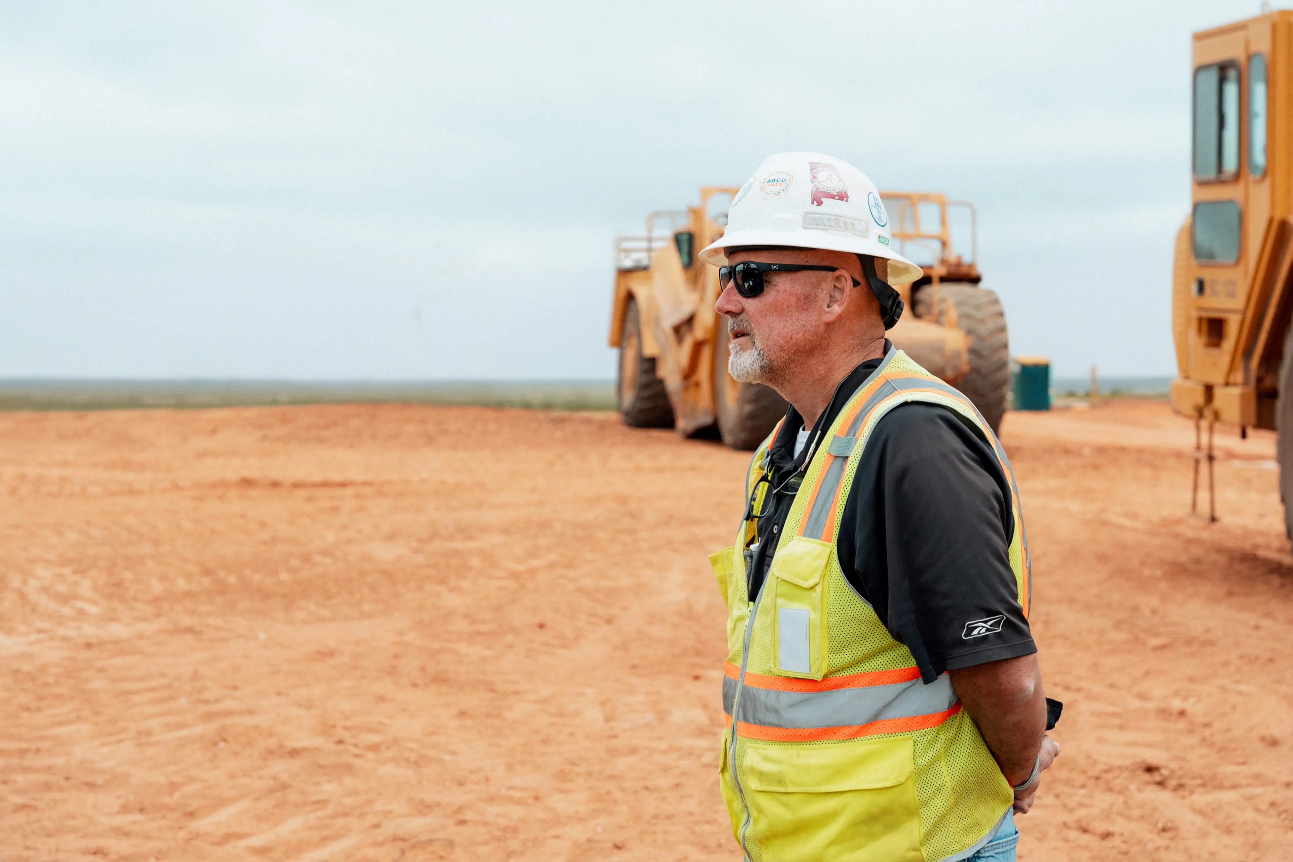 A site supervisor in a white hard hat and hi-vis vest looks across a cleared jobsite, earthmovers parked behind him